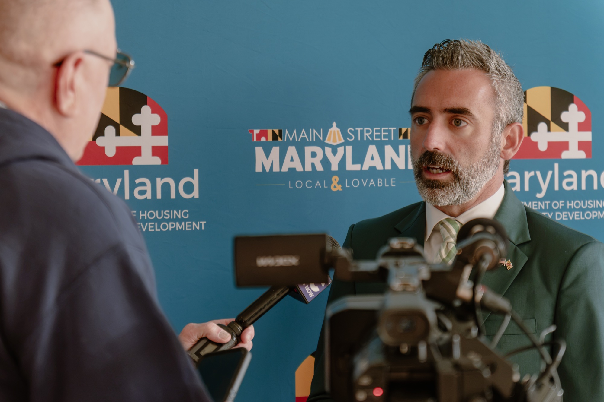 A media interview in front of the blue Main Street Maryland backdrop, with microphones and a camera in the foreground